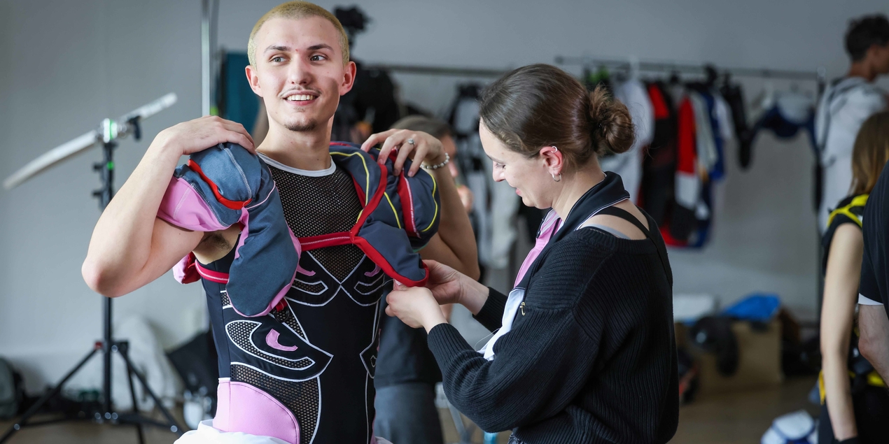 Andrej Kran trägt ein Kostüm mit Schulterpolstern und etwas, das an ein Tutu erinnert während Isabel Niemann etwas am Kostüm feststeckt