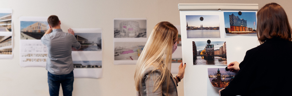 Ein Student hängt Entwürfe an einer Wand auf. Zwei Studierende schauen sich Fotos von Hamburg an.