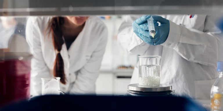 Zwei Wissenschaftlerinnen tropfen eine Flüssigkeit mit einer Spritze in ein Becherglas.