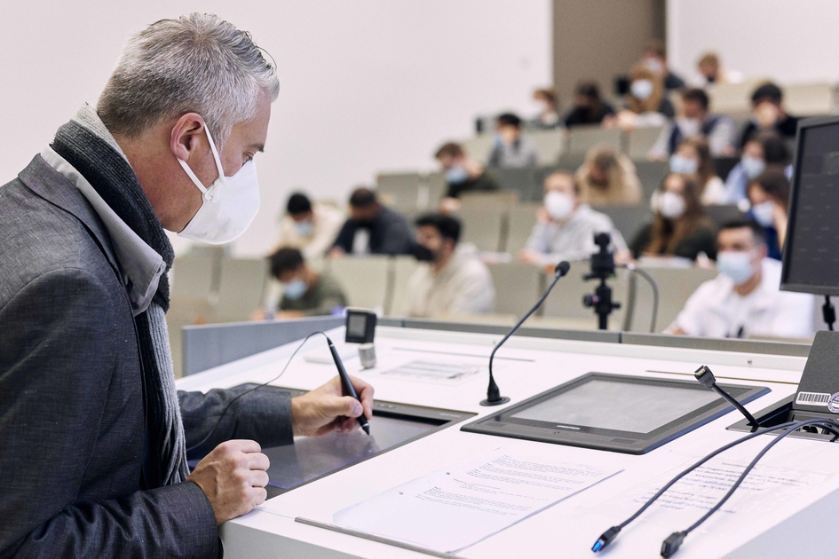 Ein Professor steht im Vordergrund an einem Rednerpult, im Hintergrund sind verschwommen Studierende im Hörsaal zu erkennen.
