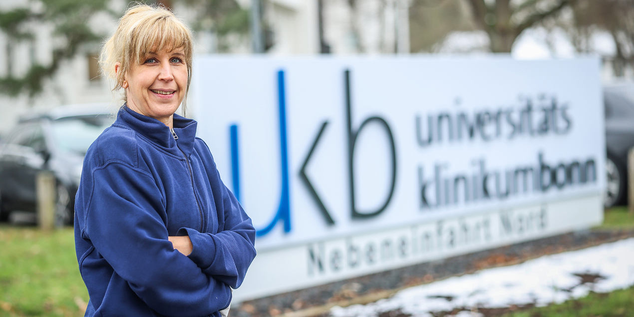 Claudia Weiß steht vor einem Logo des Universitätsklinikums Bonn.