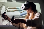 Eine Studentin trägt eine VR-Brille und hält zwei Controller in der Hand. Im Hintergrund sieht man die Übertragung des digitalen Szenarios von einer Person im Krankenbett.