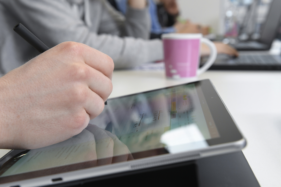 Eine Hand gibt etwas auf einem Tablet ein, im Hintergrund sind Menschen an Laptops zu erkennen