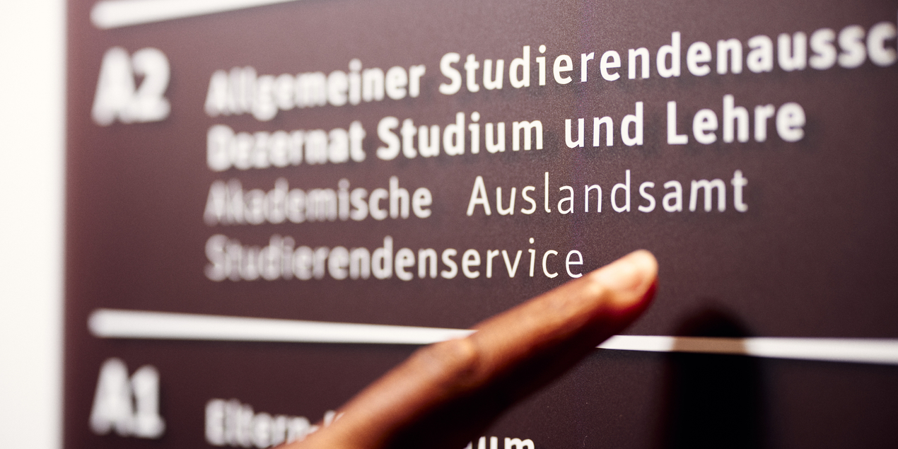 Ein Finger zeigt auf die Beschilderung des akademischen Auslandsamt in der FH Bielefeld